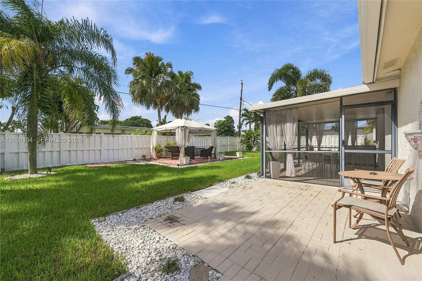 Backyard featuring a screened-in patio, concrete paver terrace, manicured lawn, white privacy fencing, and mature palm trees