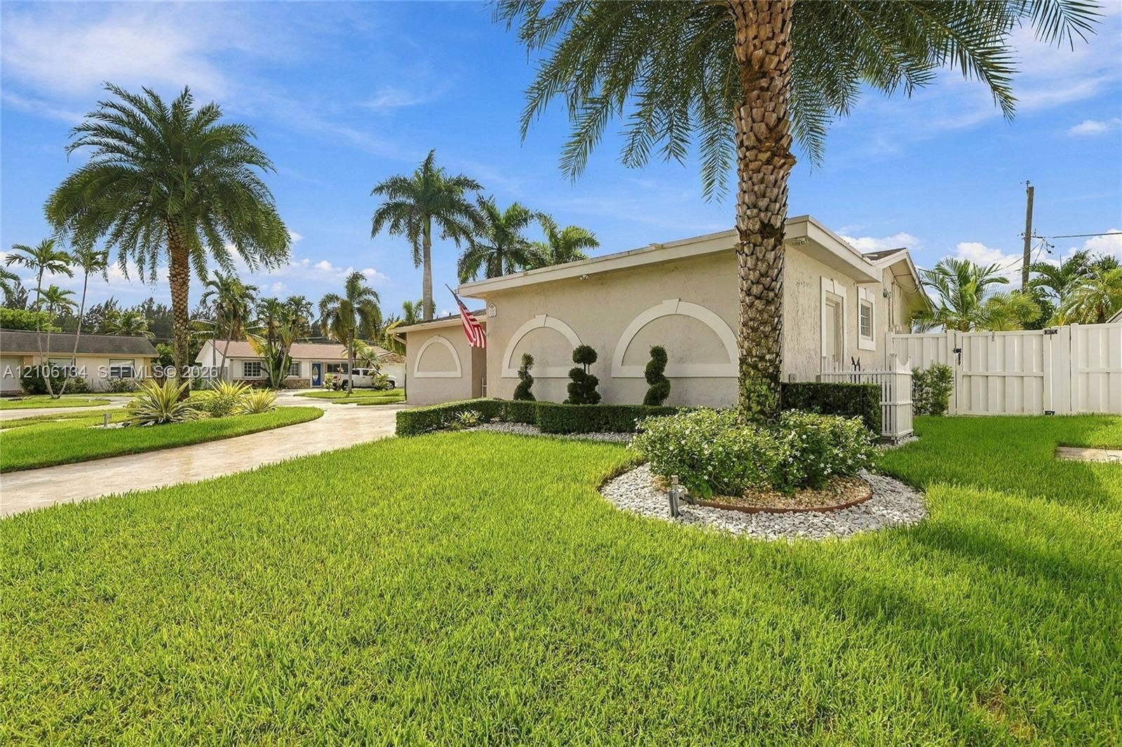 Stucco exterior featuring arched architectural accents, manicured landscaping with topiary shrubs, stone-mulched garden beds, mature palm trees, and a privacy fence