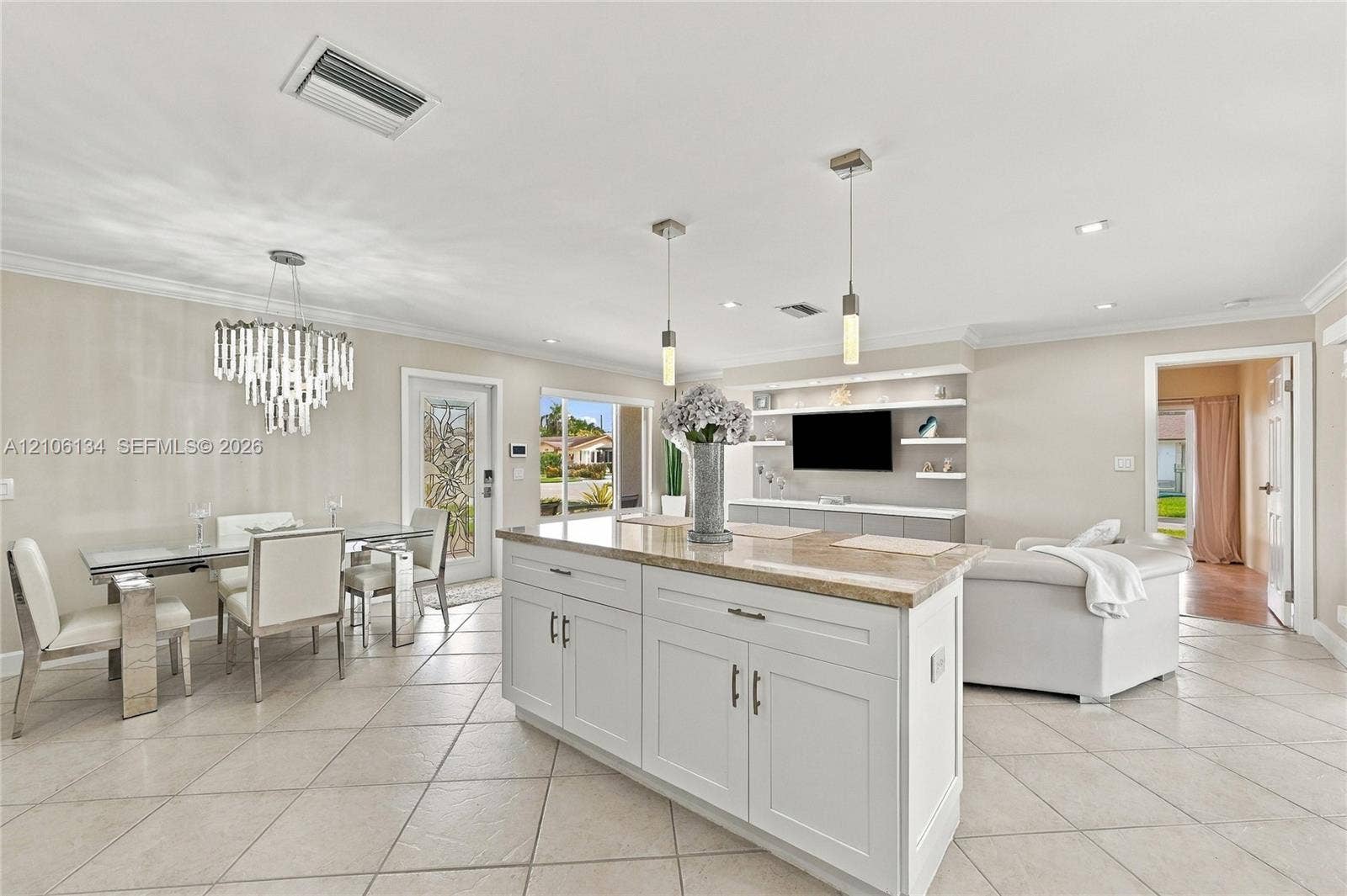 Open-concept area featuring tile flooring, a central kitchen island with light-toned countertop, custom built-in media shelving, tiered crystal chandelier, and two pendant lights