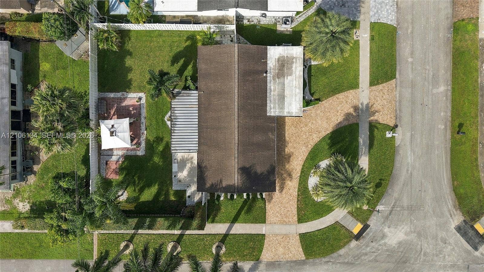 Aerial perspective of the property featuring a paved driveway, a partially covered patio area, white perimeter fencing, and established palm trees