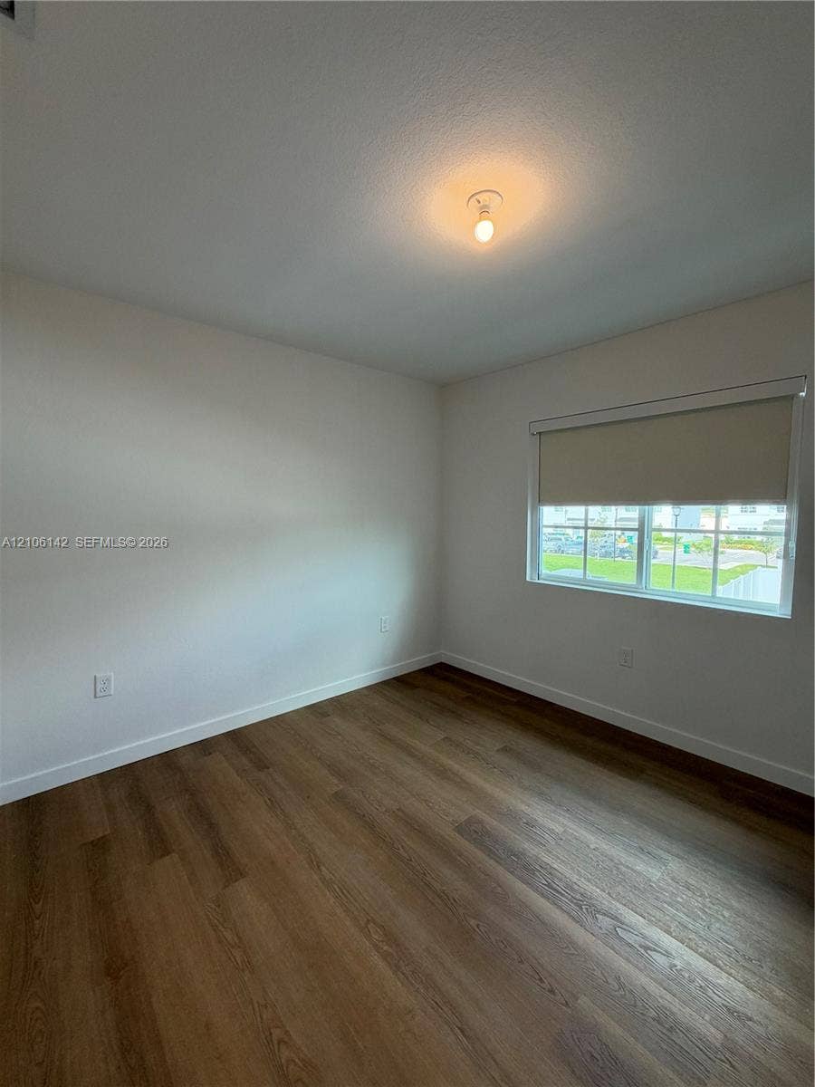 Room featuring wood-finish flooring, neutral walls, a single-pane window with a retractable shade, and a flush-mount ceiling light fixture