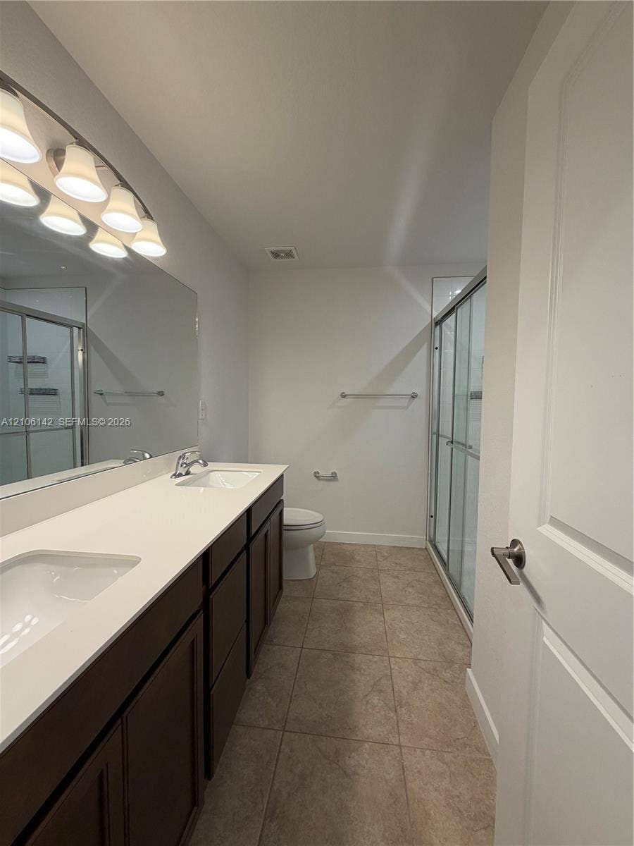 Dual-sink vanity with stone-surfaced countertop, full-width vanity mirror, tile flooring, and glass-enclosed shower stall
