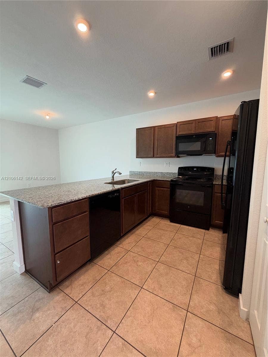 Kitchen featuring a peninsula with a stone-finish countertop, dark-toned cabinetry, ceramic tile flooring, and integrated black appliances
