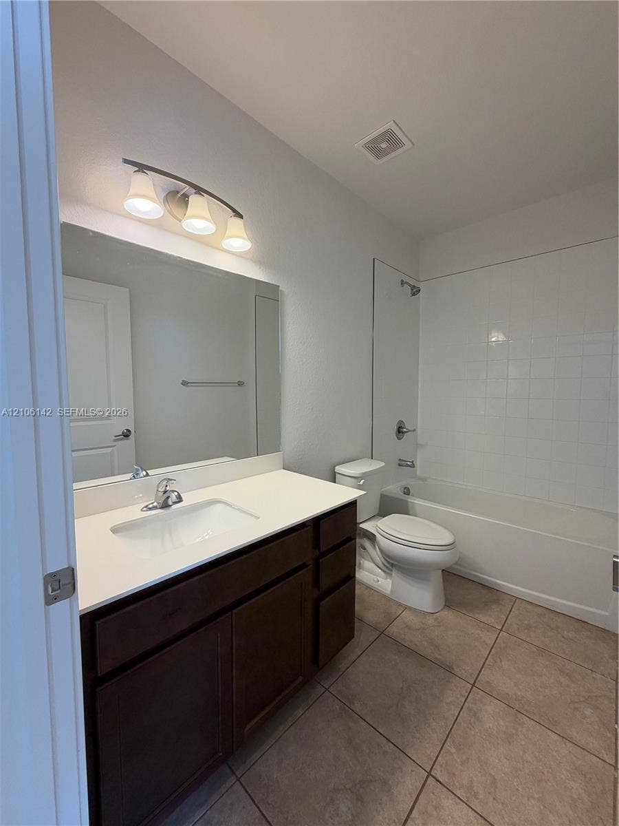 Dark wood-finish cabinetry, white countertop with integrated sink, large wall-mounted mirror, three-light vanity fixture, and tiled tub-shower surround