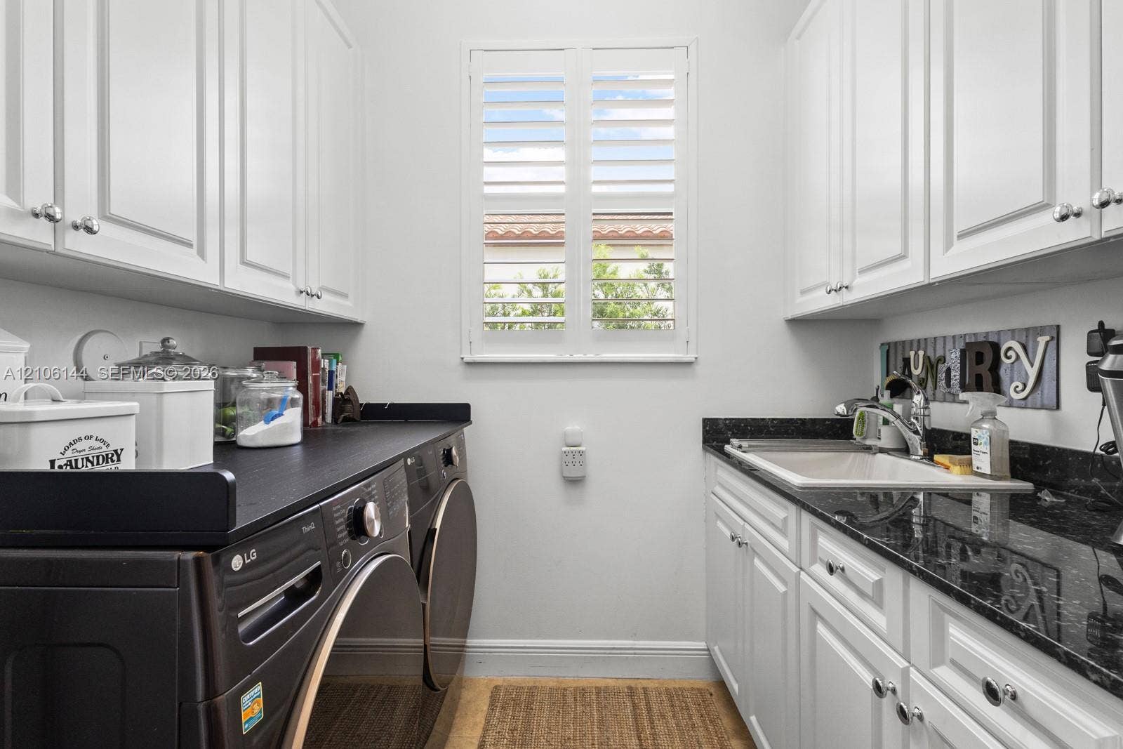 Laundry room featuring white shaker-style cabinetry, dark stone countertops, an integrated drop-in utility sink, and plantation shutters