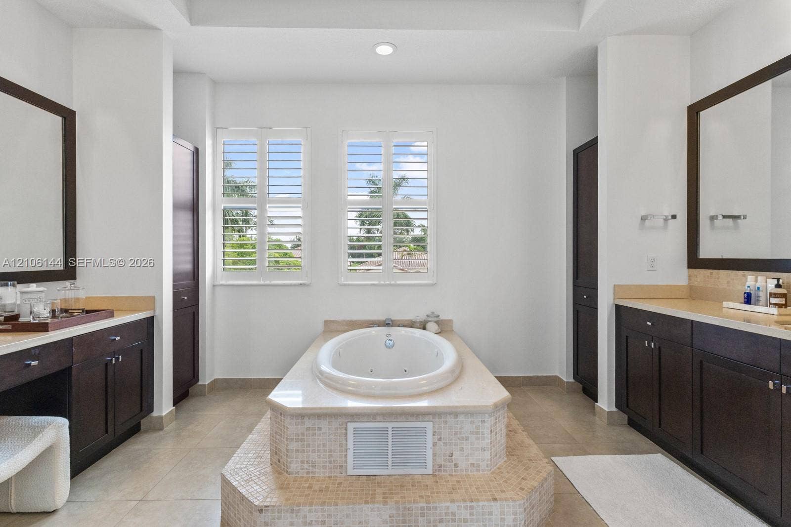 Dual vanities with dark-wood cabinetry, inset mirrors, soaking tub with a stone-surround platform, mosaic tile accents, and plantation shutters