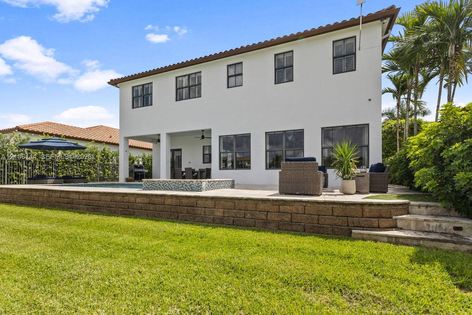 Two-story exterior featuring white stucco finish, a covered patio, a raised stone retaining wall, and an integrated pool and spa with mosaic tile accents