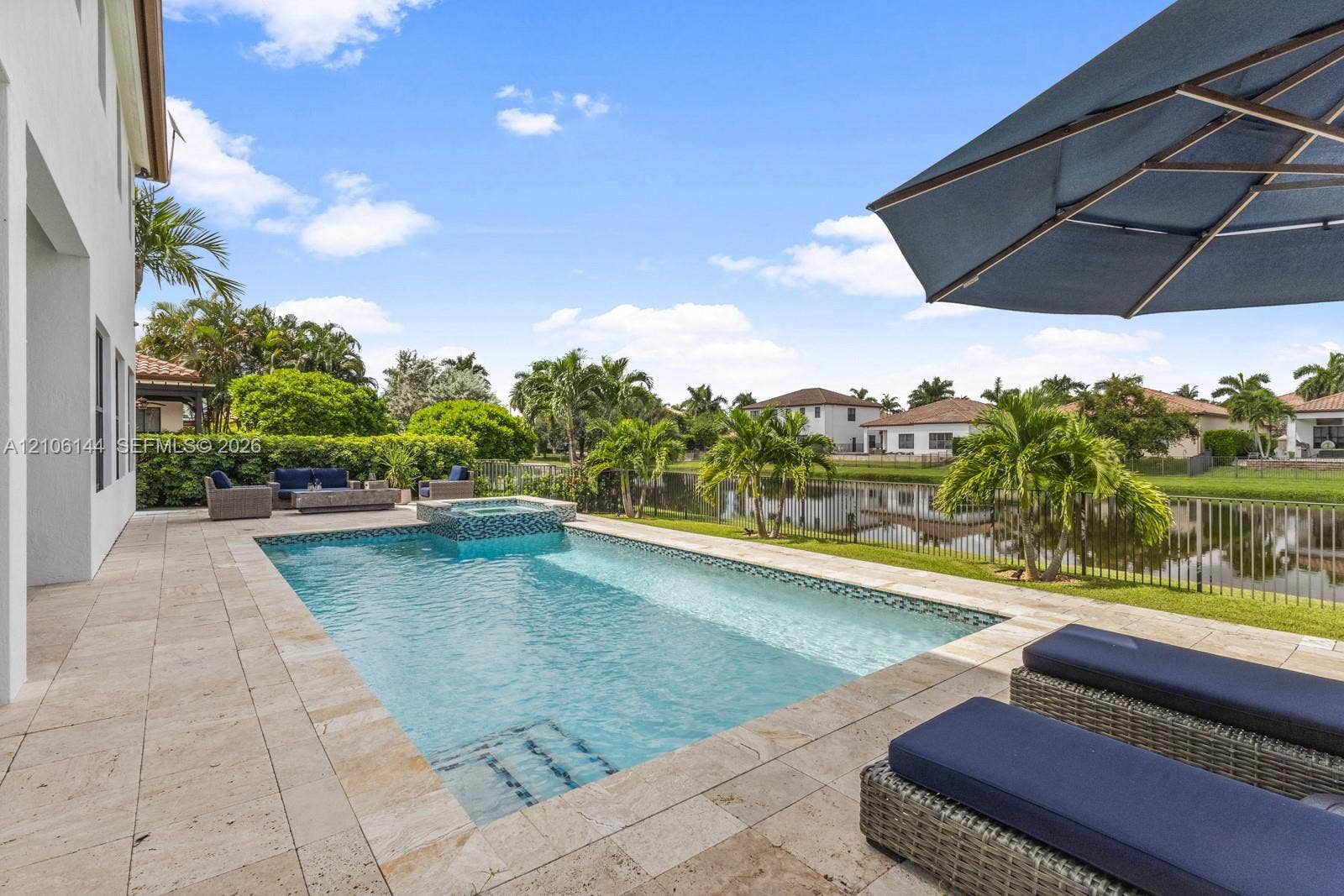 In-ground swimming pool featuring a built-in spa, travertine paver decking, and metal perimeter fencing overlooking a pond