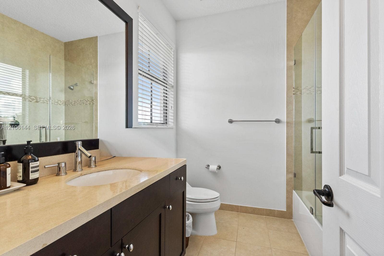 Bathroom featuring a dark wood-finish vanity, light-toned stone countertop with an undermount sink, framed wall-to-wall mirror, and a shower-tub combination with glass enclosure