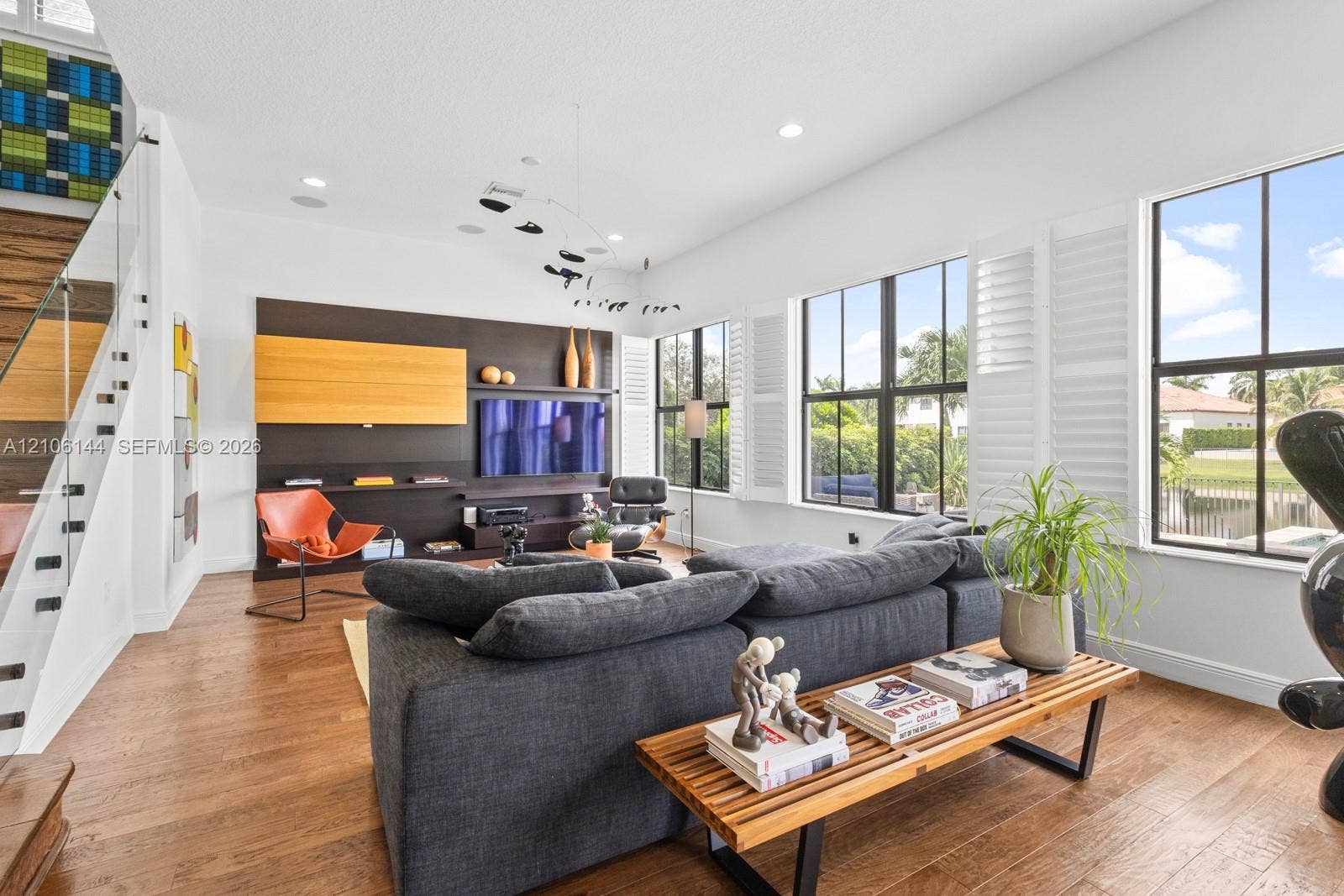 Living area featuring wood-finish flooring, floor-to-ceiling windows with plantation shutters, built-in media cabinetry, and a glass-paneled staircase railing