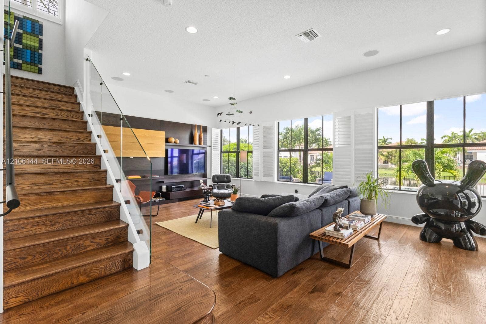 Open-concept living space featuring wood-finish flooring, a glass-panel staircase railing, built-in wall cabinetry, recessed lighting, and expansive windows with louvered shutters