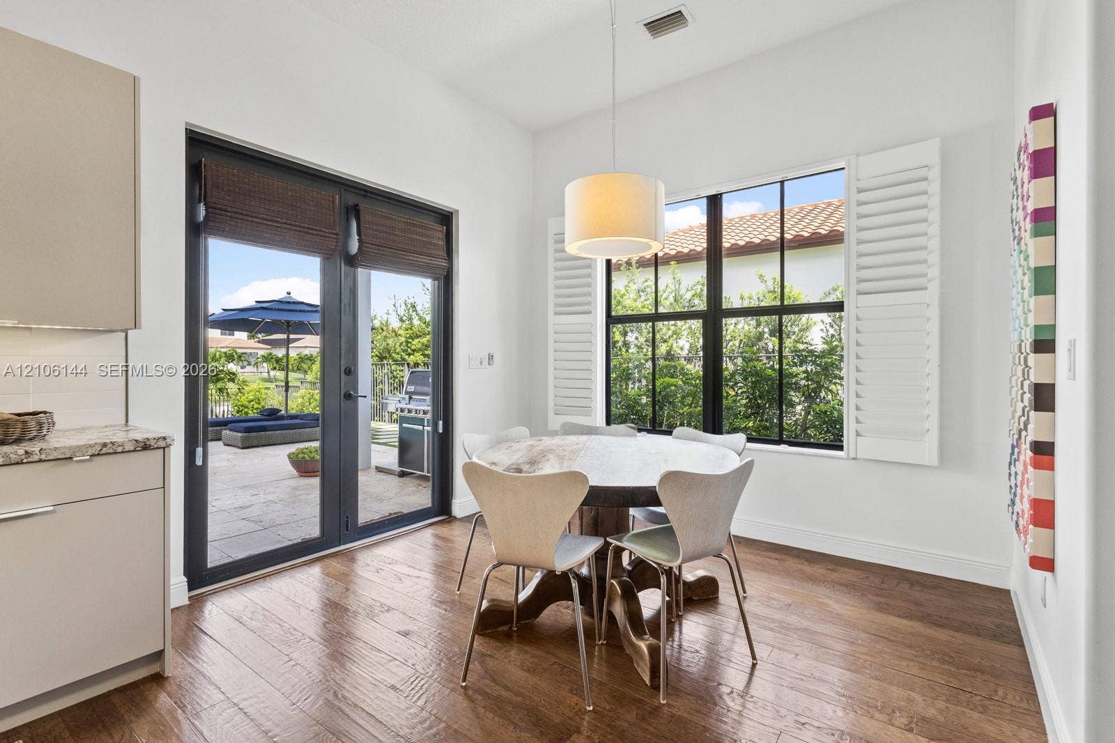 Dining area featuring wood-finish flooring, double glass doors with dark framing, multi-pane windows with white plantation shutters, and a drum-style pendant light fixture