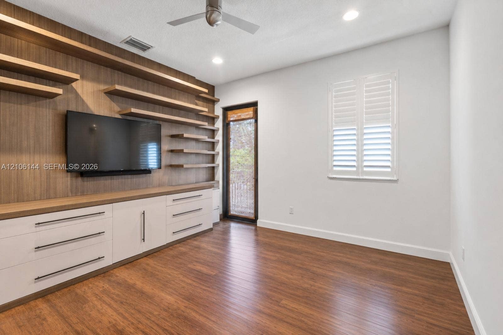 Custom floor-to-ceiling wood-finish media wall with floating shelving and integrated cabinetry, wood-finish flooring, plantation shutters, and a ceiling fan