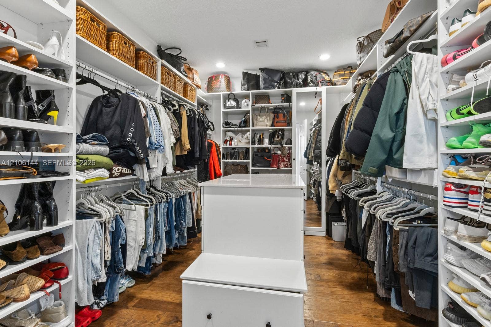 Walk-in closet featuring extensive built-in white shelving, integrated hanging rods, a central island with countertop, full-length mirror, and wood-finish flooring