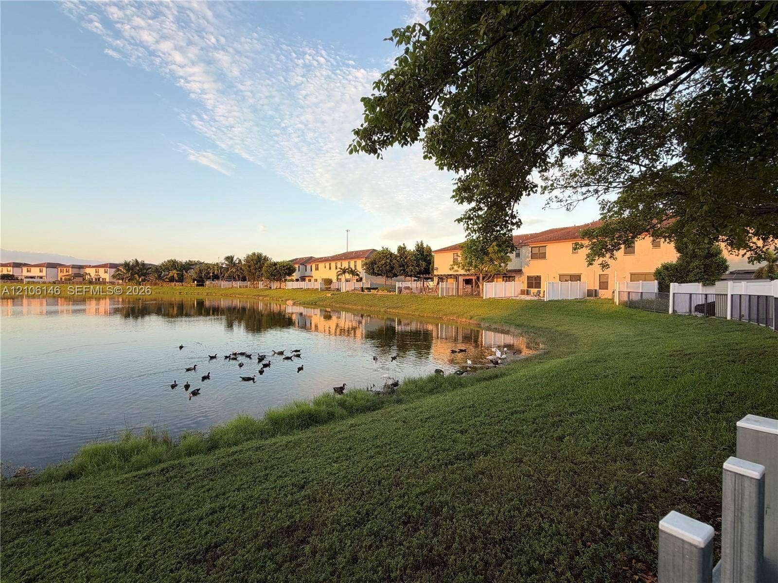 3259 W 100th Terrace, Hialeah, FL 33018 Waterfront property featuring a landscaped grass slope, mature trees, and white vinyl perimeter fencing