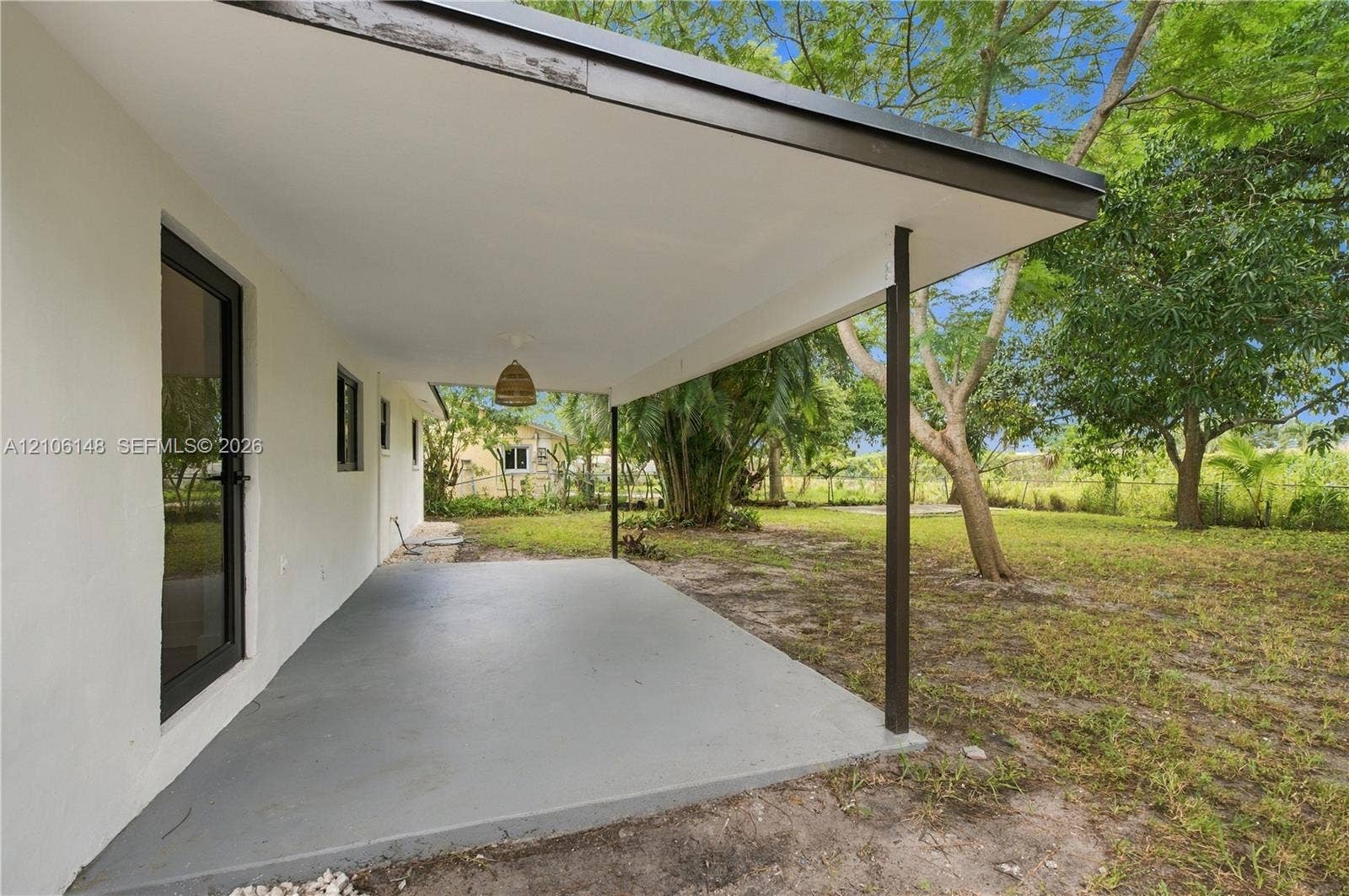 2920 Dorson Way, Delray Beach, FL 33445 Covered concrete patio featuring a solid support post, recessed ceiling lighting, and exterior access door, set against a spacious yard with mature trees