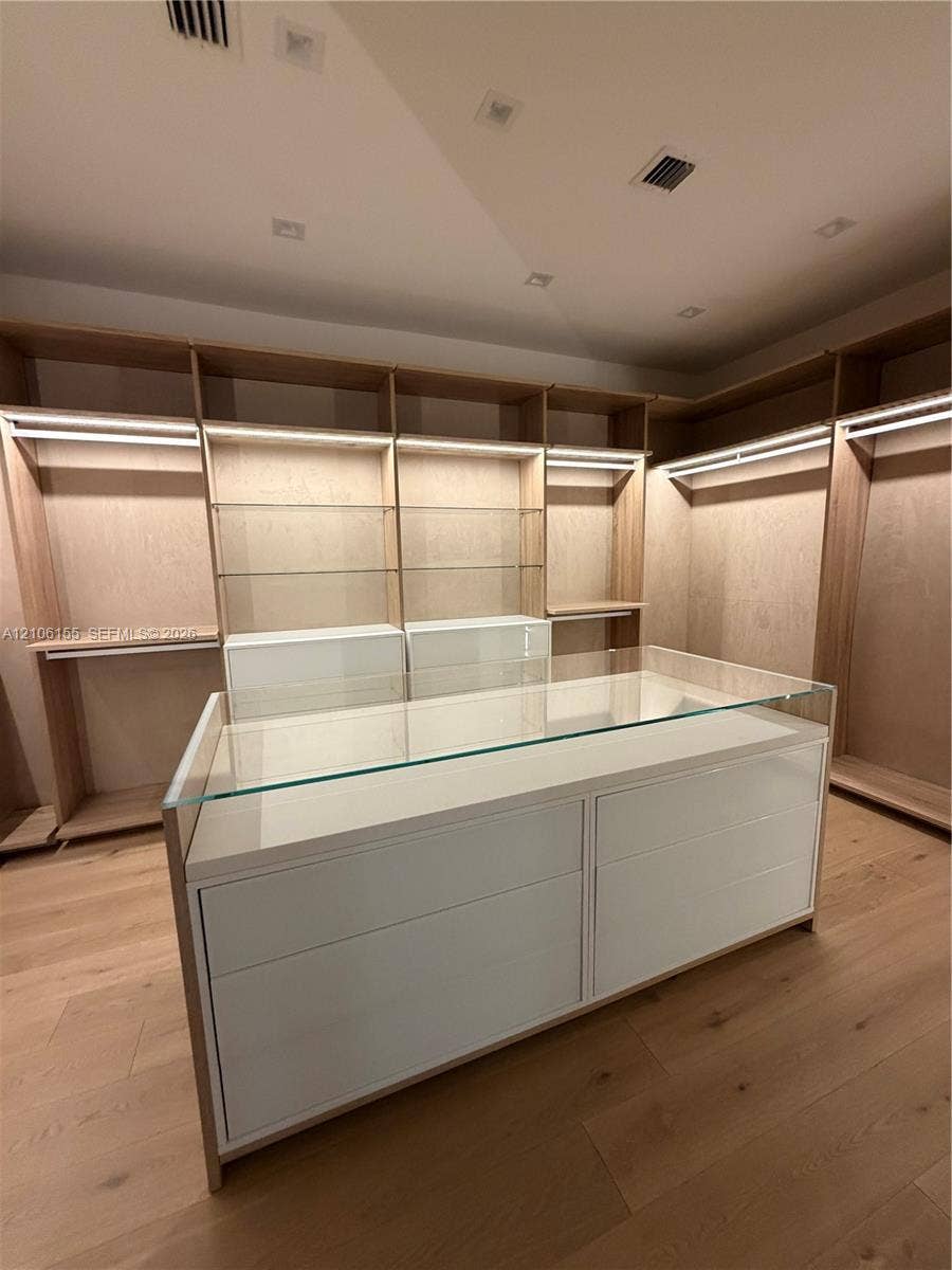 Custom Womens primary closet , long mirror ,built-in wood cabinetry featuring integrated lighting and glass shelving, centered by a matching island with a glass top and matte finish drawer fronts, complemented by wood-finish flooring and recessed ceiling lighting