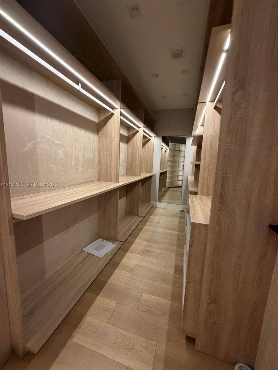 Custom Mens Primary closet w storage system featuring light wood-finish cabinetry, integrated linear LED shelving illumination, open-concept shelving units, and wood-finish flooring