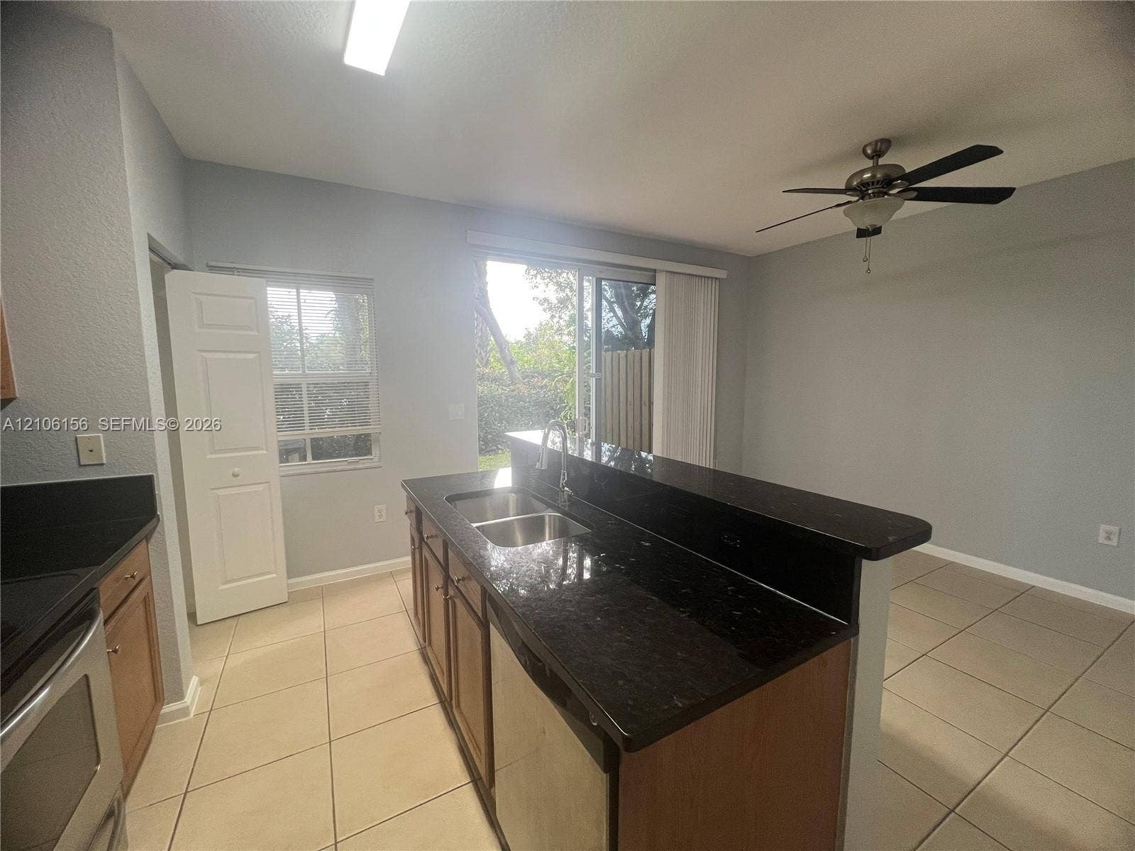 Kitchen featuring an island with a dual-basin sink, dark stone-finish countertops, wood-finish cabinetry, tile flooring, and a ceiling fan