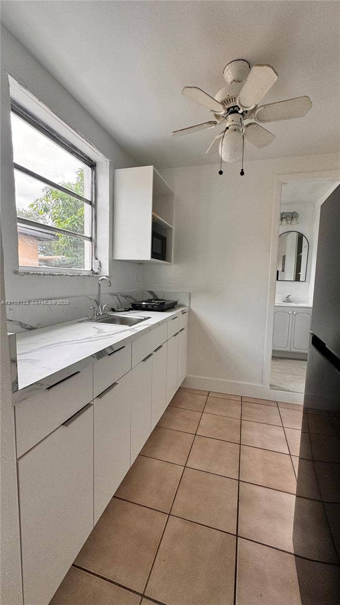 White cabinetry with minimalist hardware, stone-finish countertops, stainless steel undermount sink, multi-pane window, and ceramic tile flooring