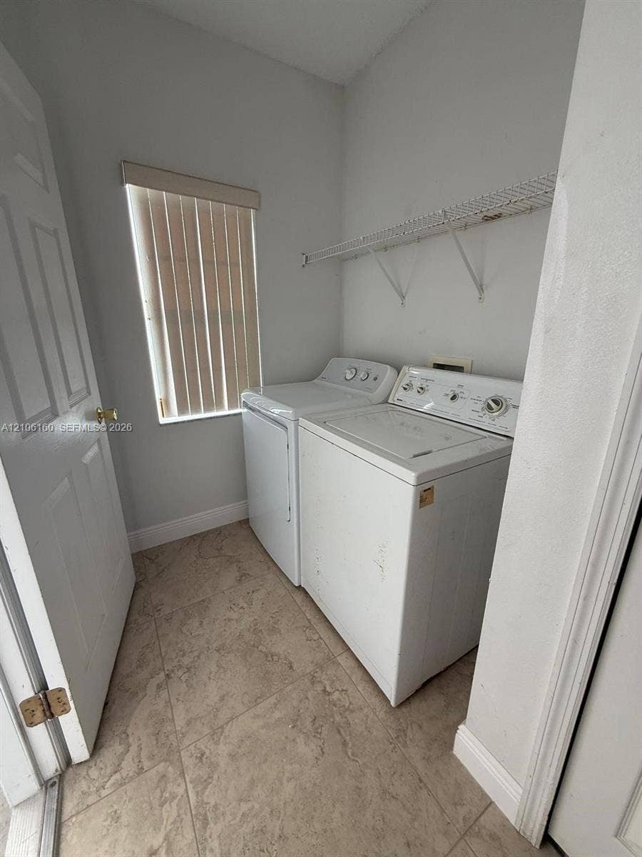 Dedicated laundry area featuring large-format tile flooring, a window with vertical blinds, and wall-mounted wire shelving