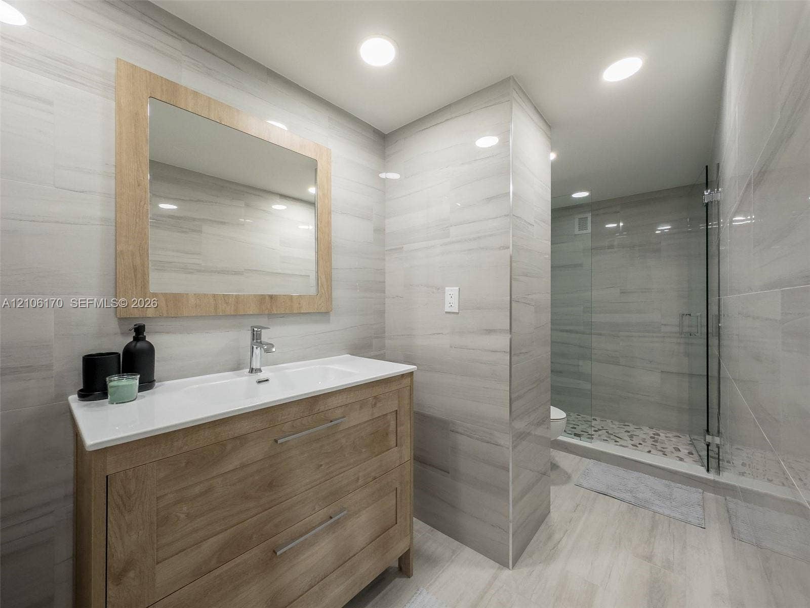 4001 S Ocean Drive, #12B, Hollywood, Hollywood, FL 33019 Wood-finish vanity with a rectangular sink, integrated mirror with wood frame, recessed lighting, and a glass-enclosed shower featuring a pebbled floor