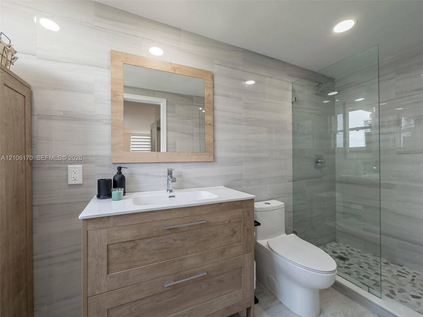 4001 S Ocean Drive, #12B, Hollywood, Hollywood, FL 33019 Bathroom vanity with wood-finish cabinetry, white countertop, integrated basin, and matching framed mirror, complemented by floor-to-ceiling tile walls and a glass-enclosed shower with pebble-stone flooring