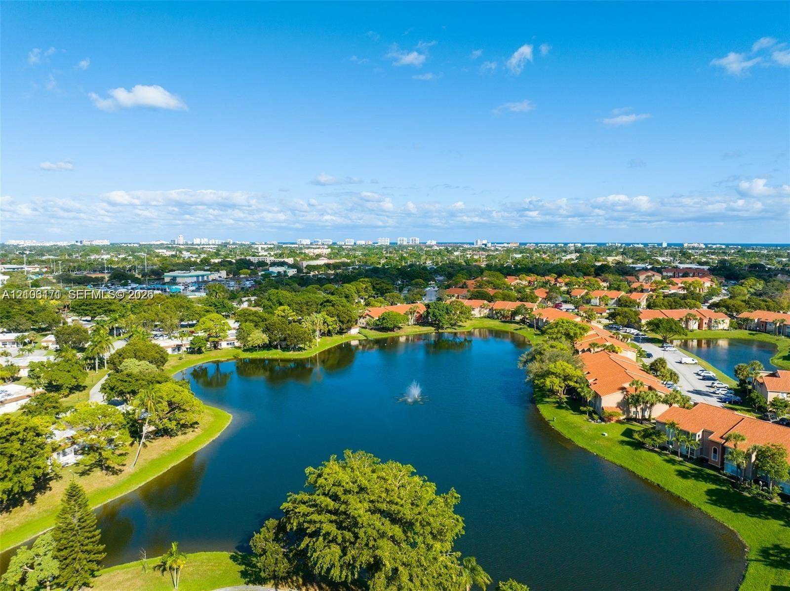Aerial perspective of a lakefront community featuring a central fountain, manicured green space, mature trees, and tile-roof building architecture
