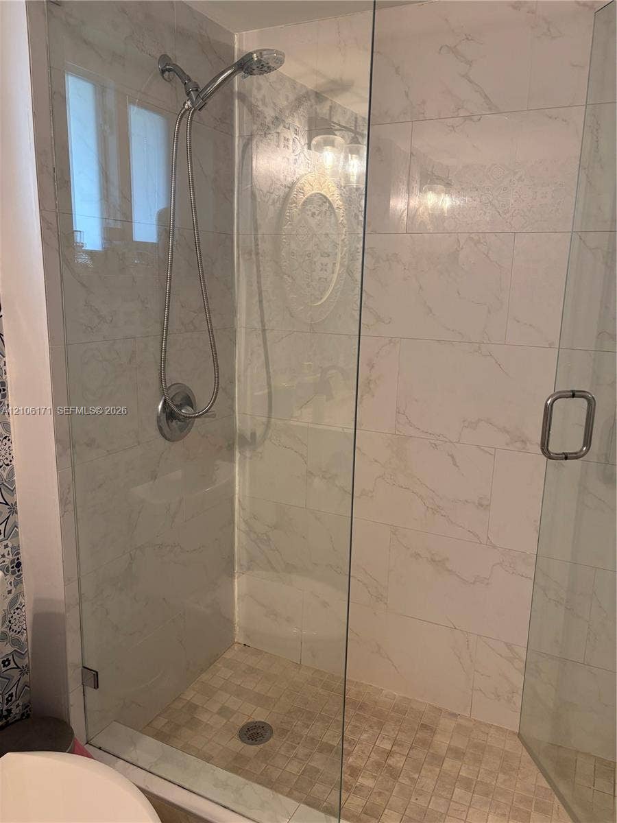 Walk-in shower featuring large-format stone-look wall tiling, a glass enclosure with a metallic handle, a pebble-finish floor, and a polished chrome handheld shower fixture