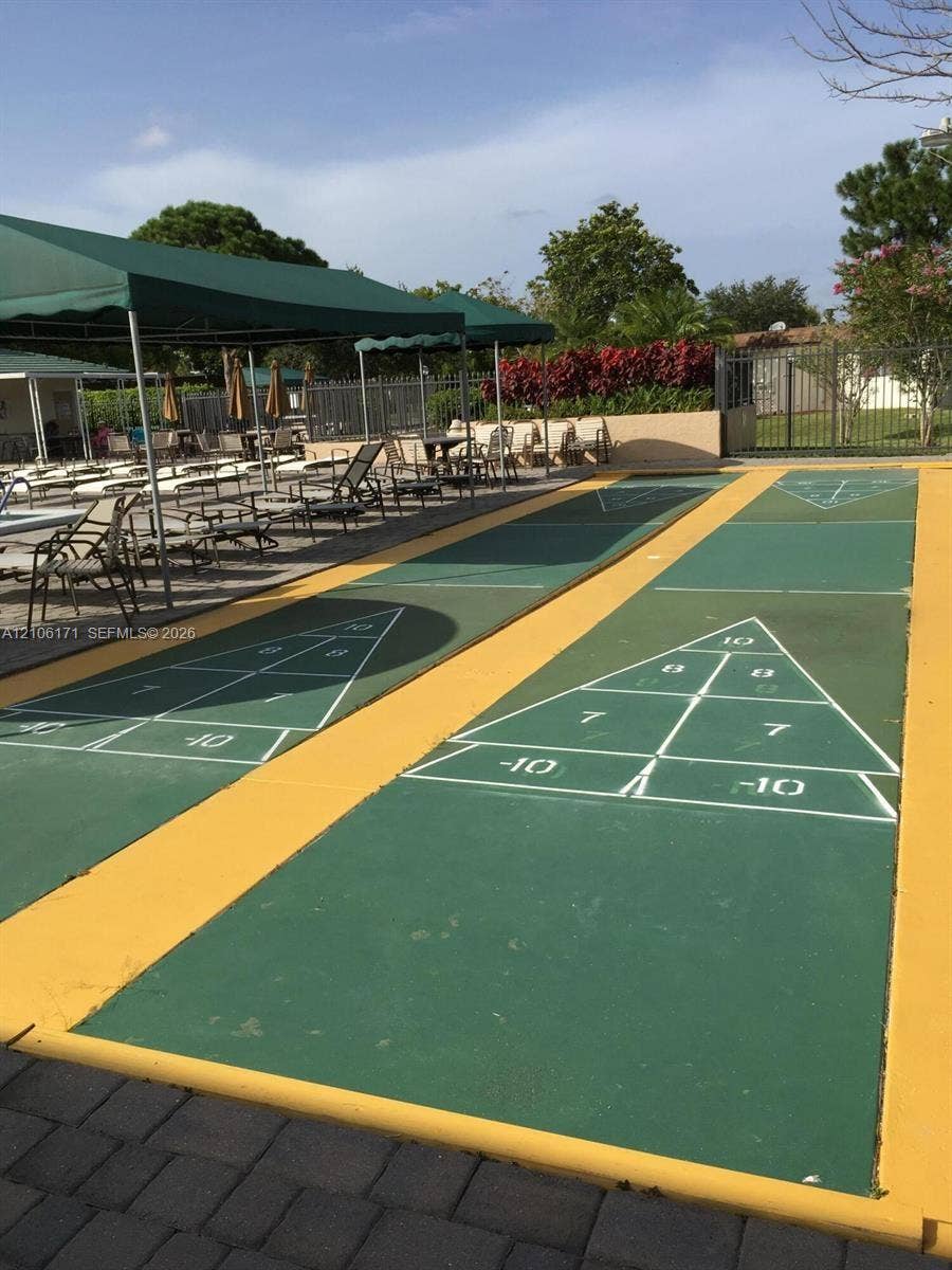 Dual shuffleboard courts featuring a green-painted surface with white regulation markings and yellow-painted borders, complemented by a covered patio area with metal-frame green awnings