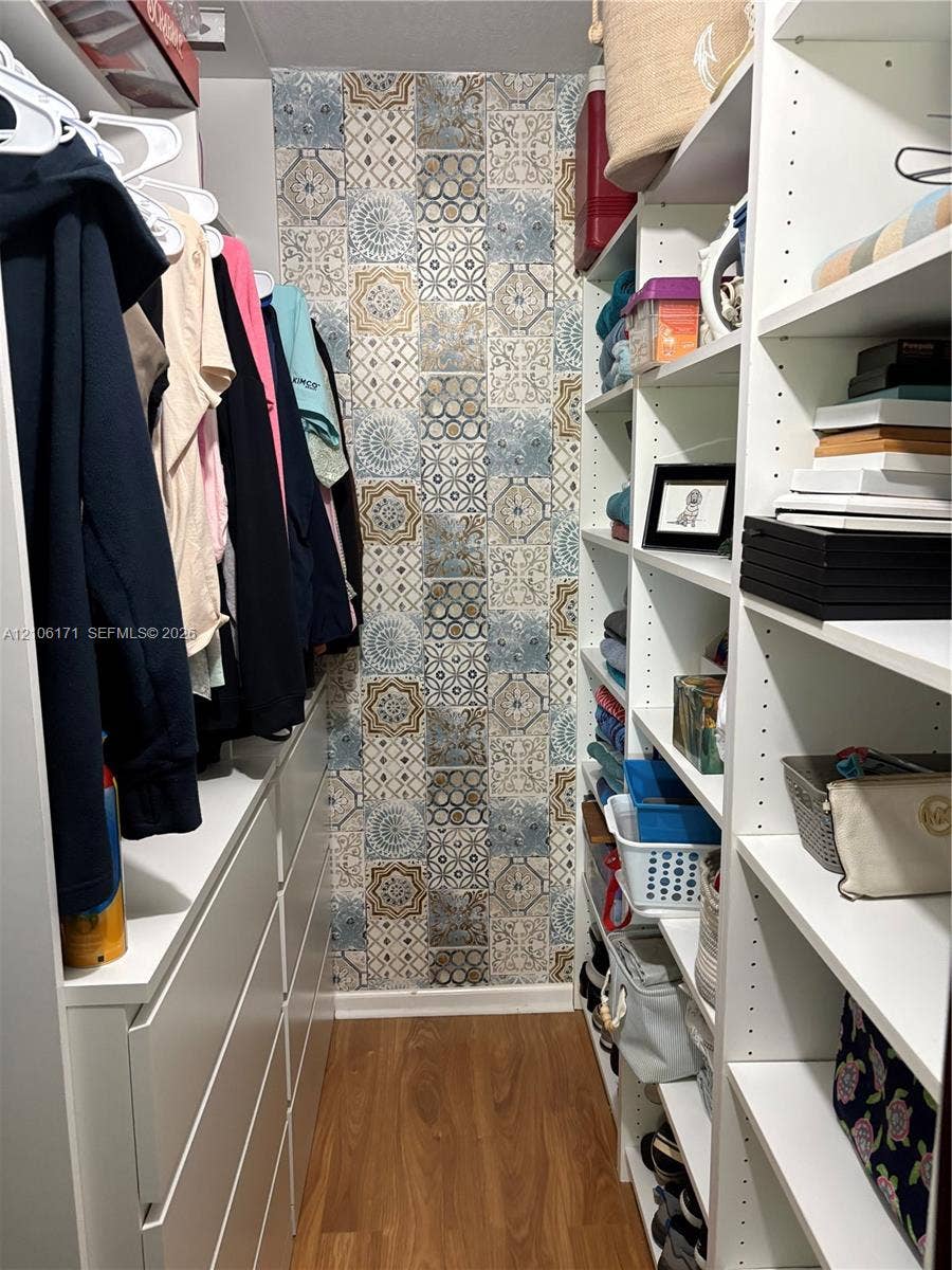 Walk-in closet featuring white built-in shelving, integrated dresser drawers, patterned accent wall covering, and wood-finish flooring