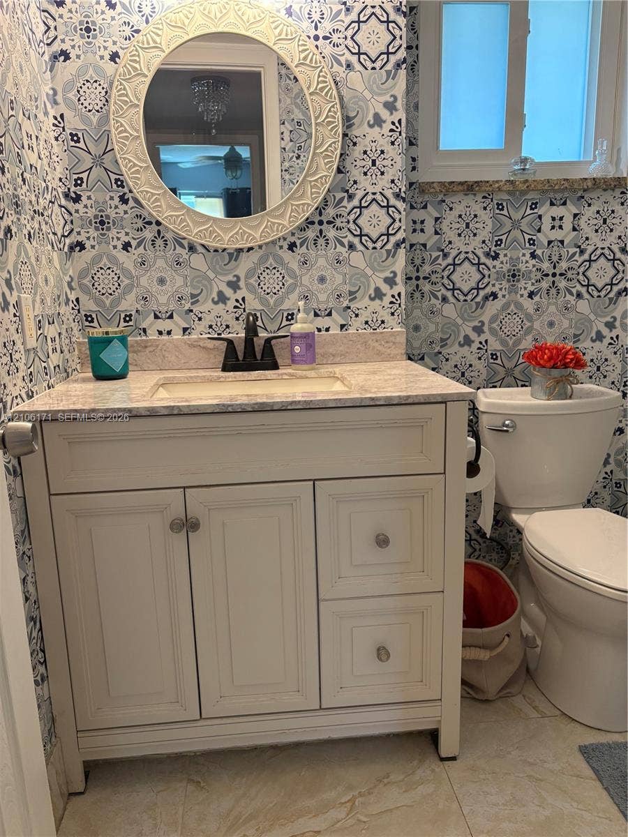 Bathroom featuring decorative patterned wall tile, a white vanity with a neutral-toned stone countertop, an integrated sink, a dark finish centerset faucet, and a circular wall-mounted mirror