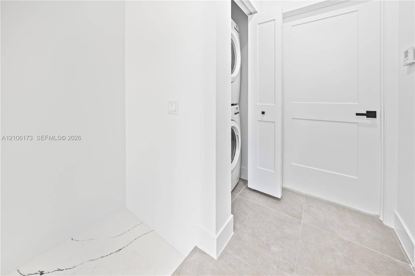 Laundry area featuring a stacked washer and dryer, large-format tile flooring, a solid-core door with matte black hardware, and stone-finish built-in counter surface