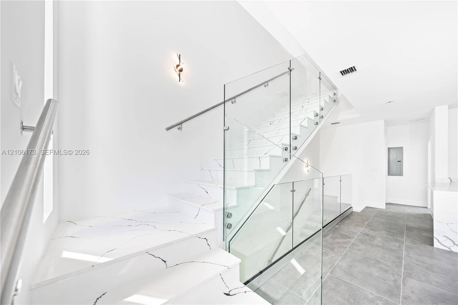 Staircase featuring white treads with veined pattern, frameless glass panel railing, and brushed metal wall-mounted handrail, complemented by large-format grey tile flooring