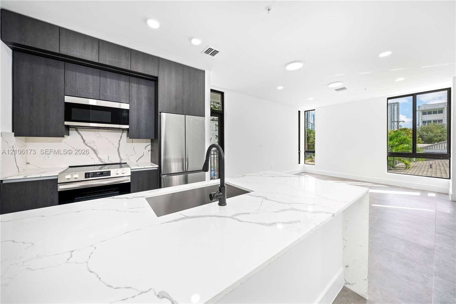Kitchen featuring matte dark cabinetry, expansive stone-finish waterfall island, stainless steel appliances, patterned stone-finish backsplash, and recessed lighting