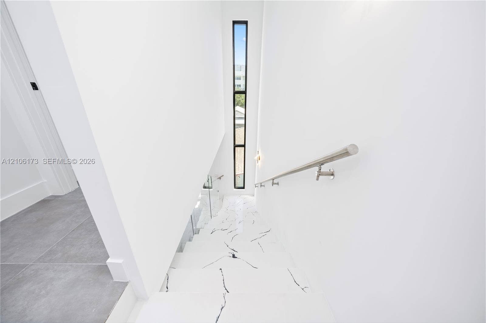 Staircase featuring white stone treads with grey veining, a stainless steel wall-mounted handrail, full-height vertical window, and glass panel railing