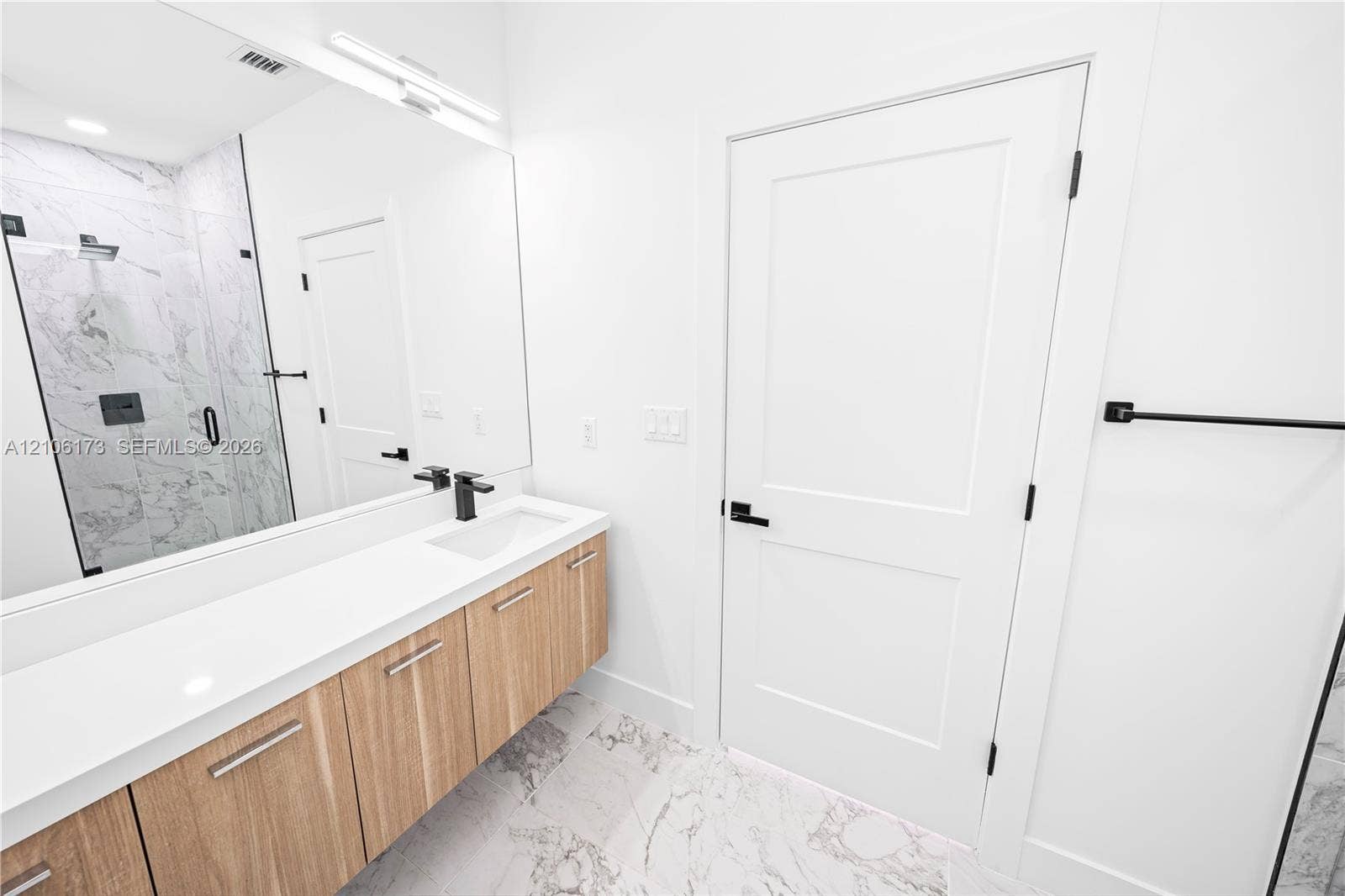 Floating wood-finish vanity with a white countertop and rectangular undermount sink, matte black faucet, glass-enclosed shower with tiled surround, and large-format tile flooring