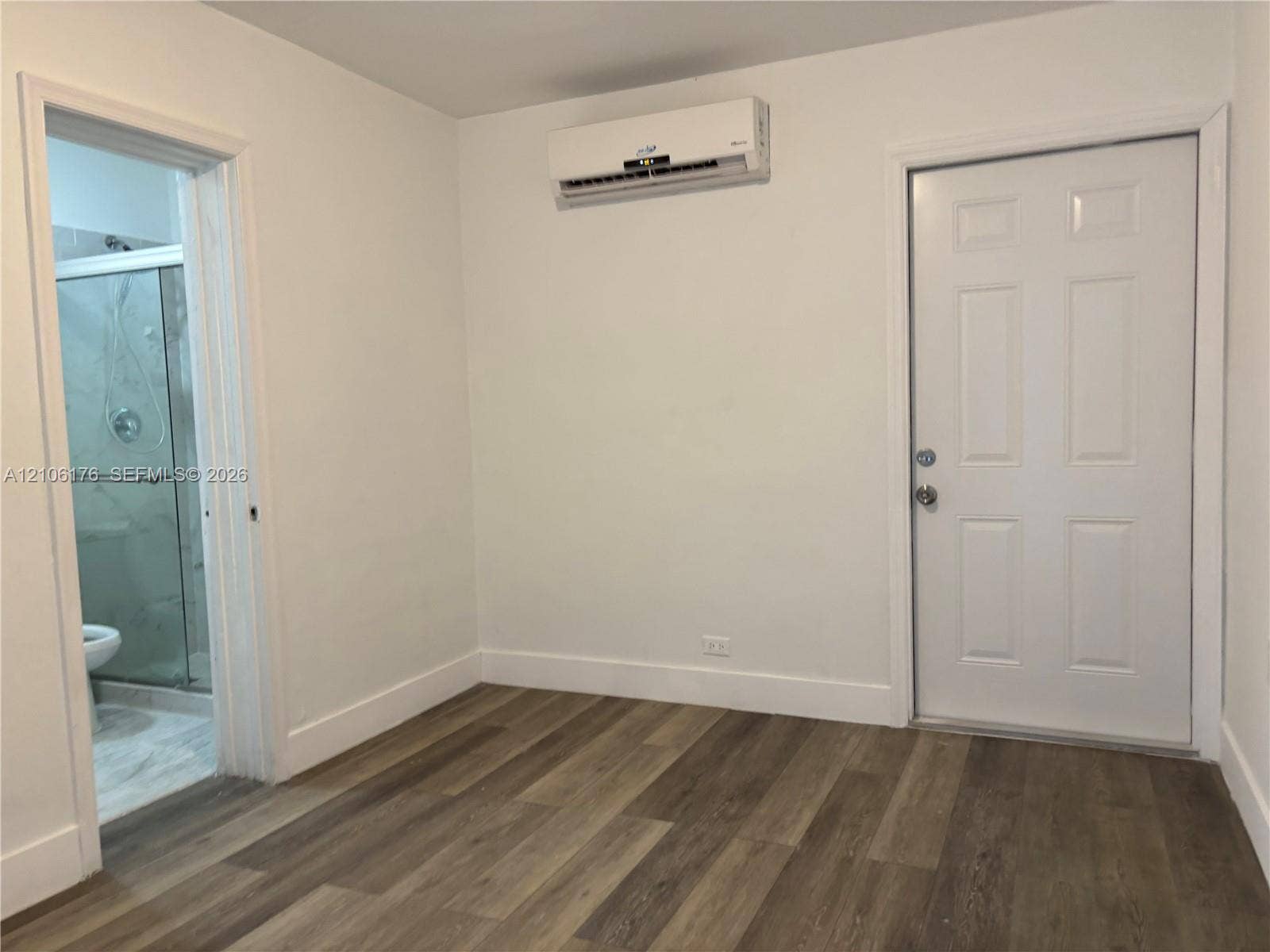 214 NW 46th Avenue, #214, Miami, Miami, FL 33126 Room featuring wood-finish flooring, a wall-mounted ductless air conditioning unit, and a six-panel door, with a glass-enclosed shower stall visible through the doorway