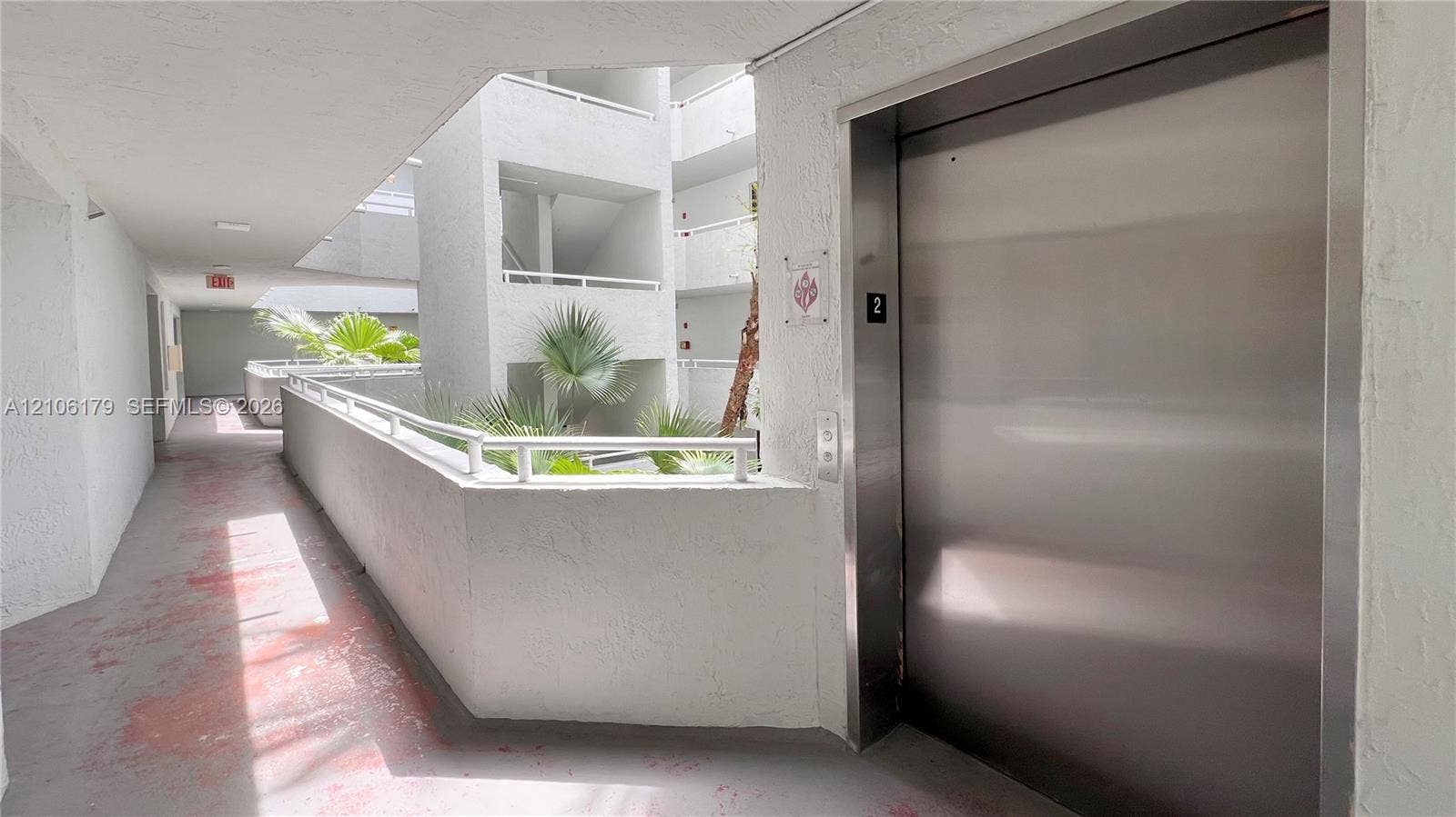 Stainless steel elevator door with minimalist metal hardware, flanked by a stucco-finished exterior hallway and white metal railings overlooking a landscaped central atrium