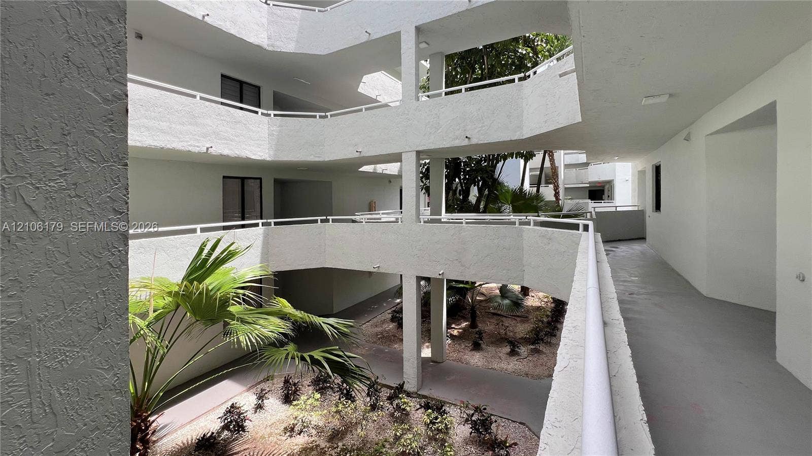 Multi-story open-air atrium featuring stucco wall finishes, metal railings, and a central landscaped courtyard with palm trees