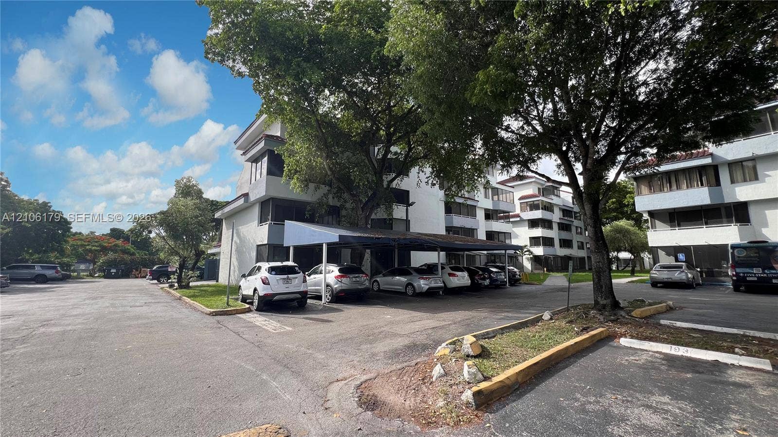 Multi-story building exterior featuring covered parking structures, paved surface parking, and mature trees