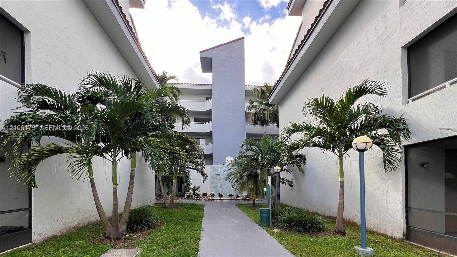 Multi-story stucco exterior featuring a paved walkway, mature palm trees, manicured landscaping, and post-mounted outdoor lighting