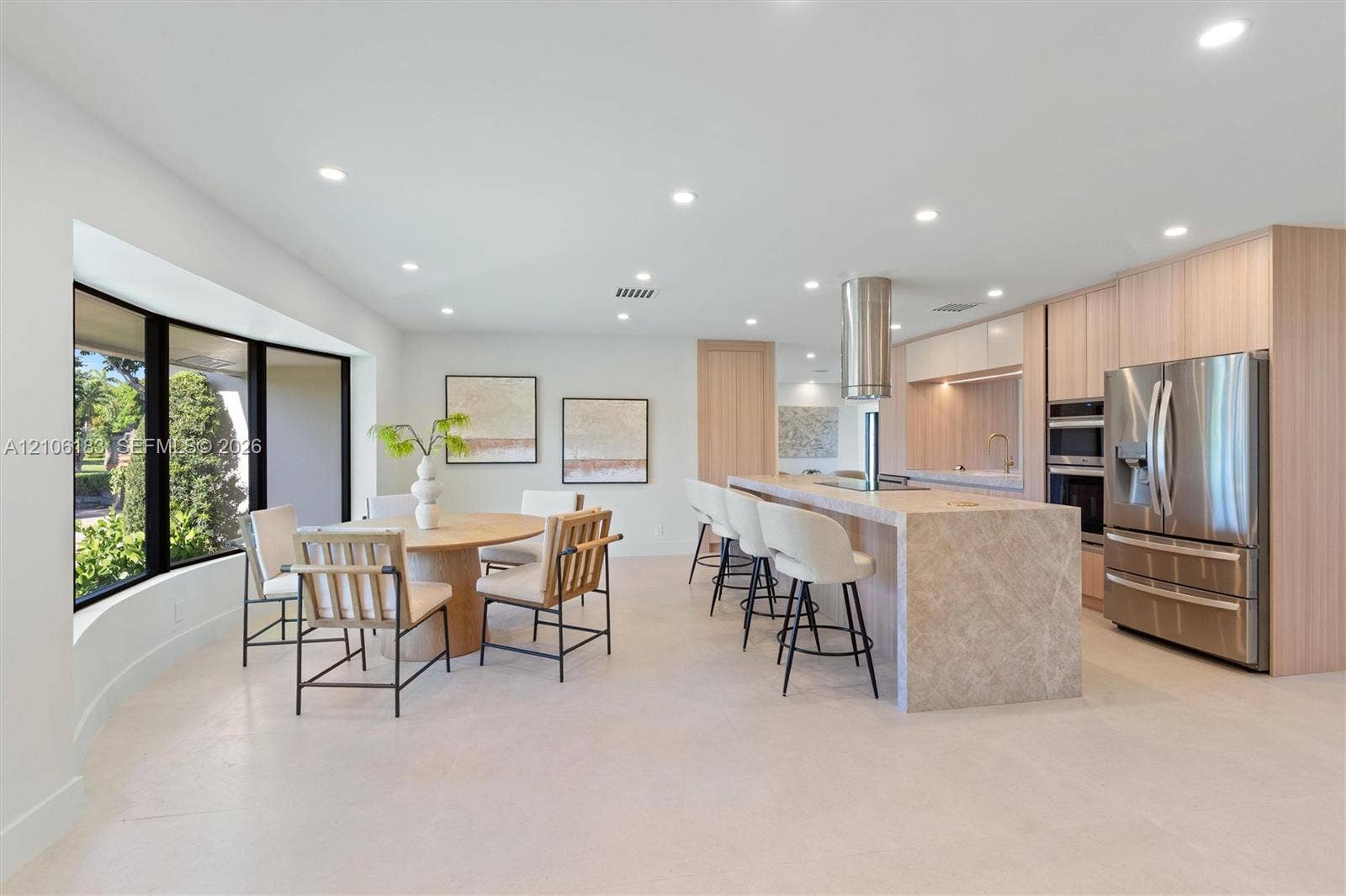 Modern kitchen featuring light wood-finish cabinetry, a large stone-surfaced island with a cylindrical exhaust hood, stainless steel appliances, recessed ceiling lighting, and expansive floor-to-ceiling windows