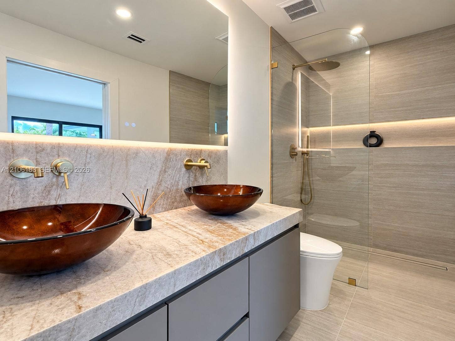 Bathroom vanity featuring a stone countertop, dual glass vessel sinks, wall-mounted gold-tone faucets, and a walk-in shower with floor-to-ceiling tile and integrated lighting