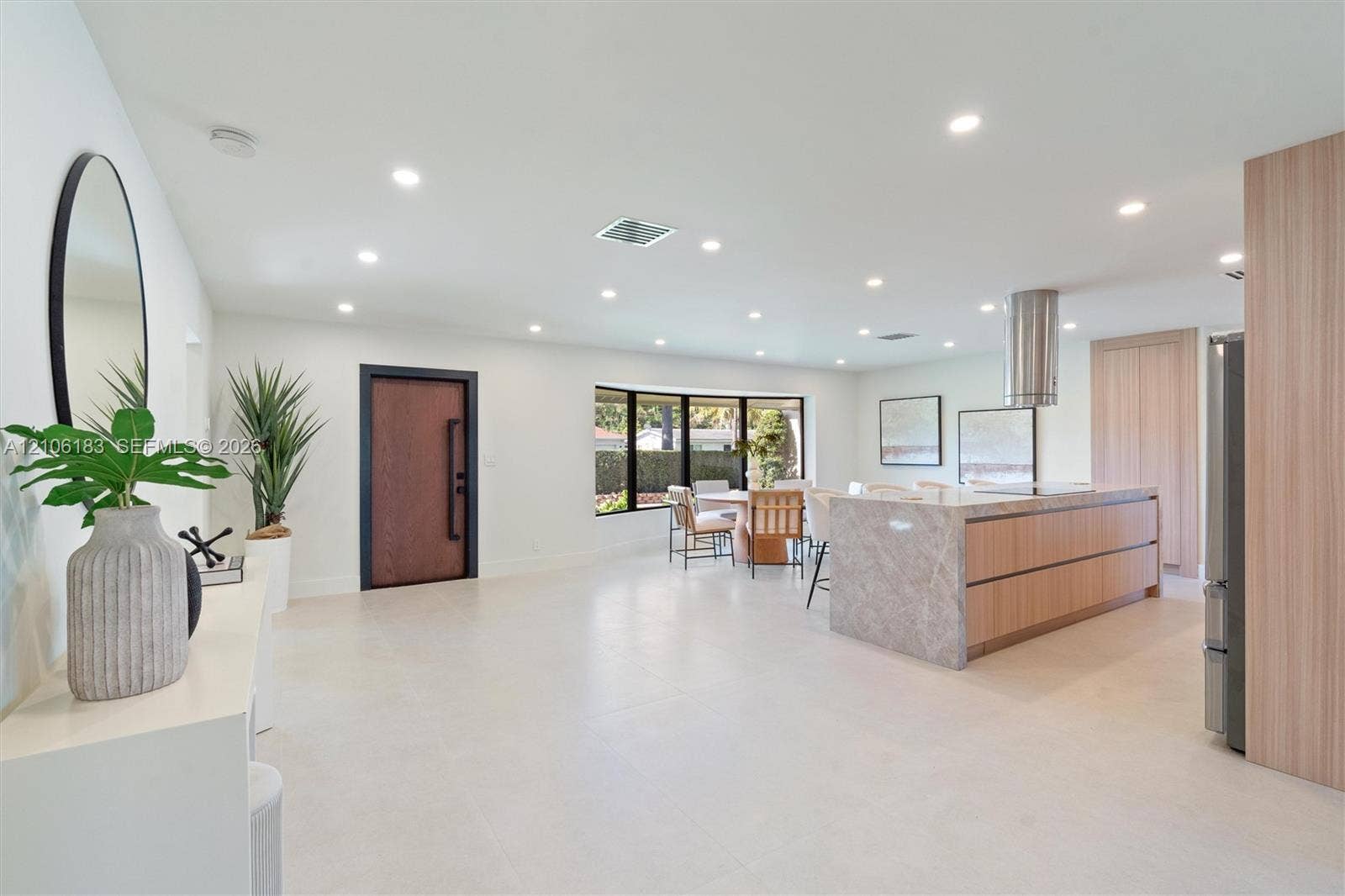 Open-concept space featuring large-format tile flooring, recessed ceiling lighting, a waterfall-edge kitchen island, natural wood-finish cabinetry, and a black-framed front door