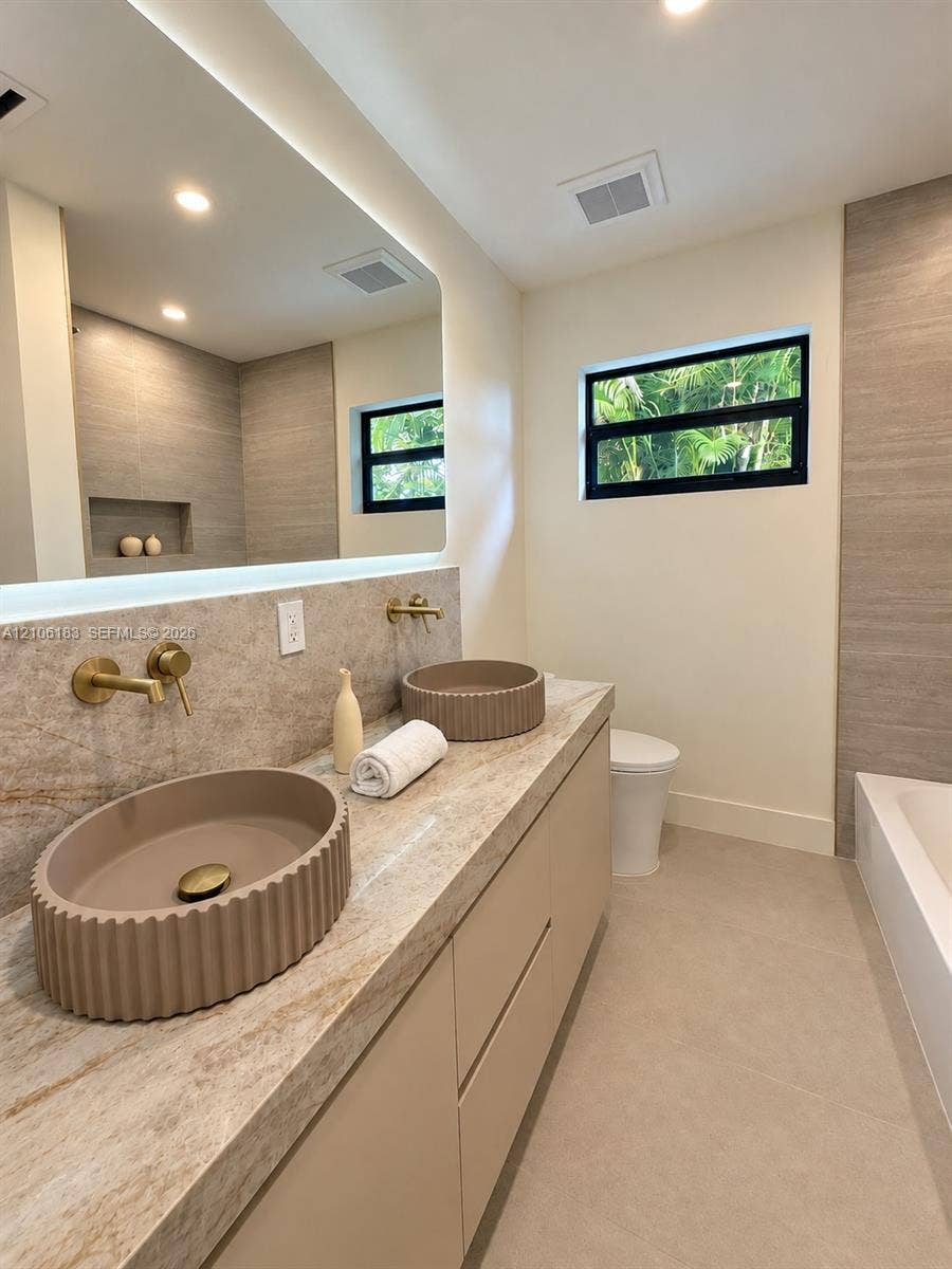 Floating vanity with dual fluted vessel sinks, stone-finish countertop, wall-mounted brass faucets, back-lit vanity mirror, and large-format tile wall accents