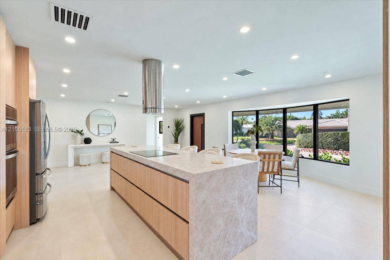 Kitchen featuring a central island with a waterfall-edge stone countertop, cylindrical stainless steel range hood, built-in wall oven, flat-panel cabinetry, and large-format tile flooring