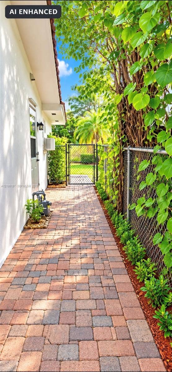 Side yard featuring a multi-tone paver walkway, metal chain-link fencing, a metal gate, and exterior wall-mounted lighting