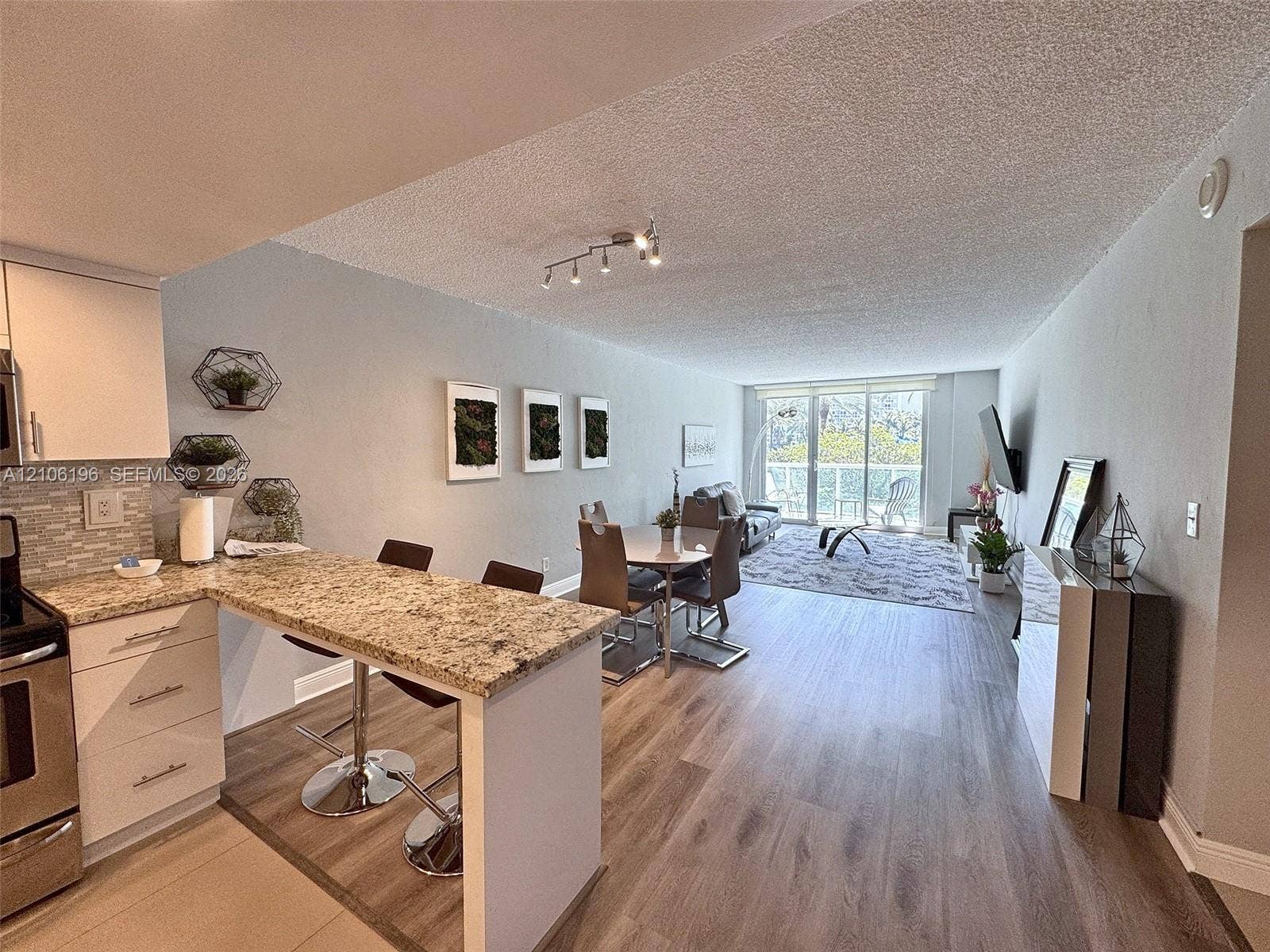 Open-concept living space featuring wood-finish flooring, stone-surfaced breakfast bar, stainless steel appliances, track lighting, and floor-to-ceiling glass sliding doors