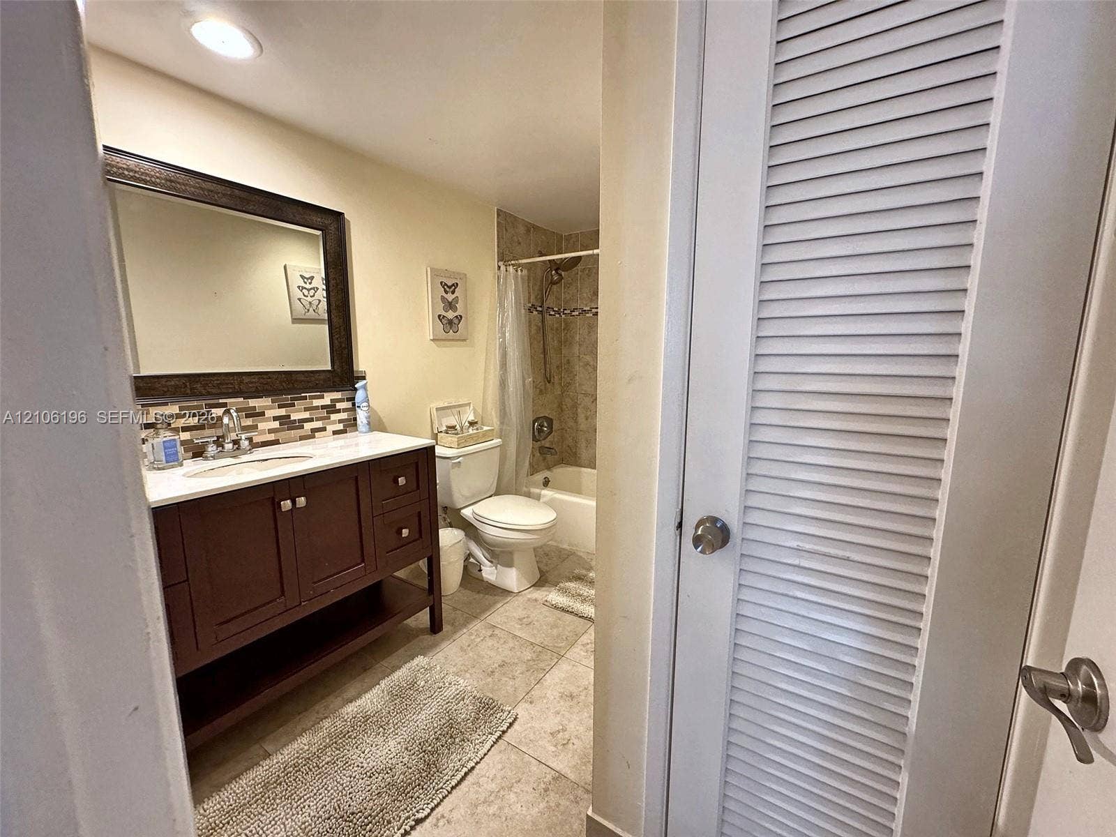 Bathroom featuring a dark wood vanity with a light-toned countertop, multi-colored tile backsplash, framed mirror, and a closet with a louvered door